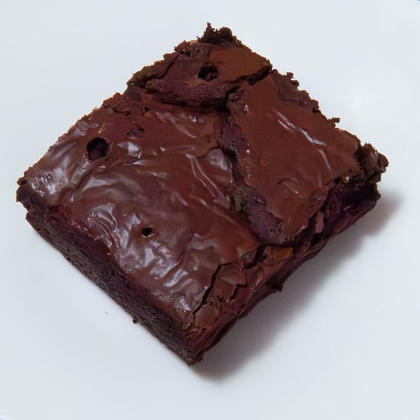 A rich, dark chocolate brownie with a shiny, crinkled top sits on a white surface.